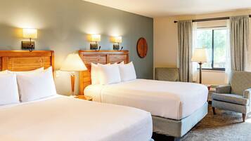 A hotel room with two beds, a wooden headboard, a nightstand, a lamp, a chair, and a window with curtains.