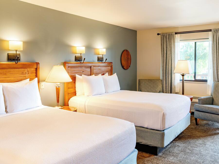 A hotel room with two beds, a wooden headboard, a nightstand, a lamp, a chair, and a window with curtains.