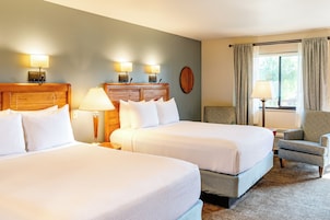Habitación de hotel con dos camas, cabecero de madera, mesita de noche, lámpara, silla y ventana con cortinas.