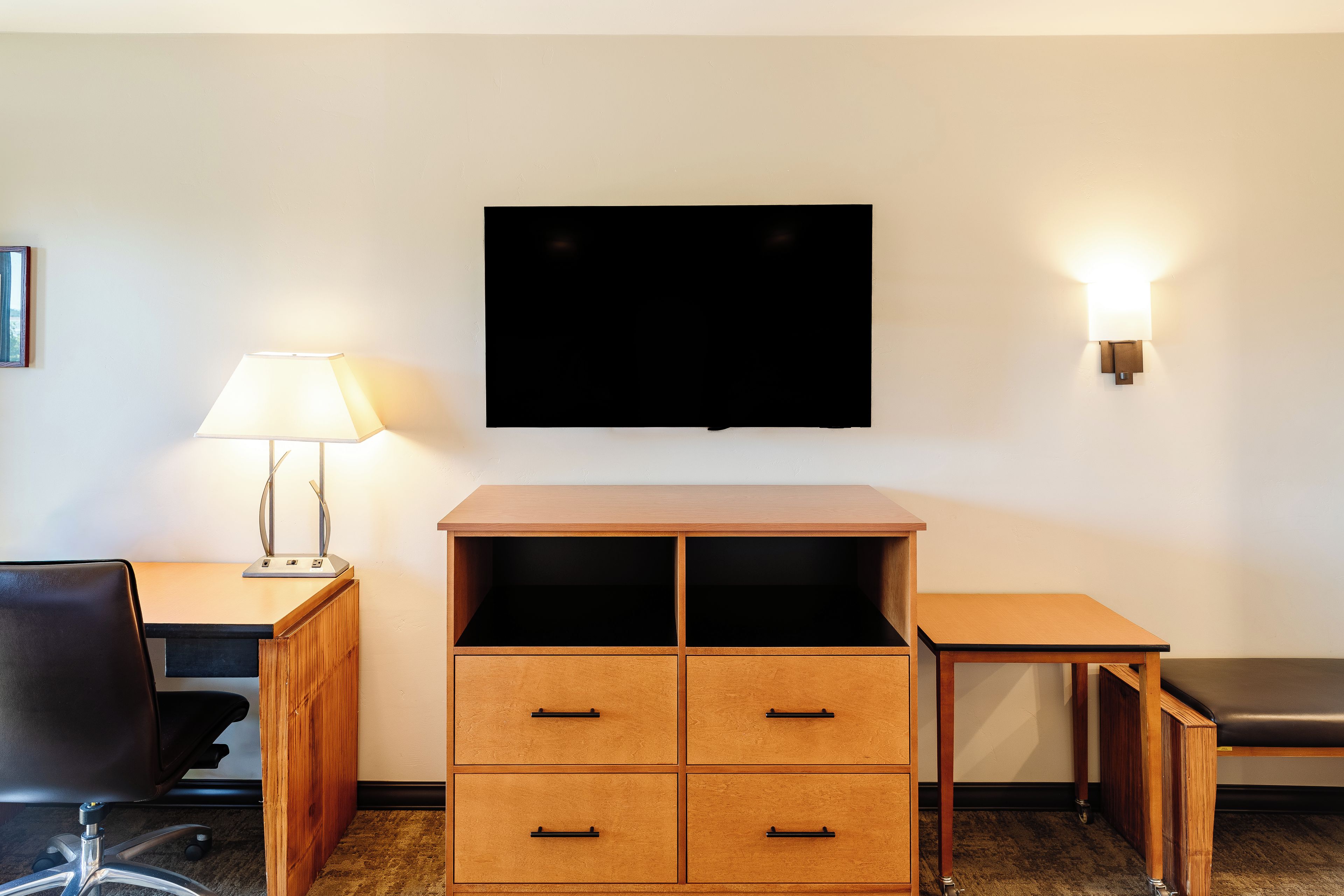 A hotel room with a desk, chair, TV, and a lamp.