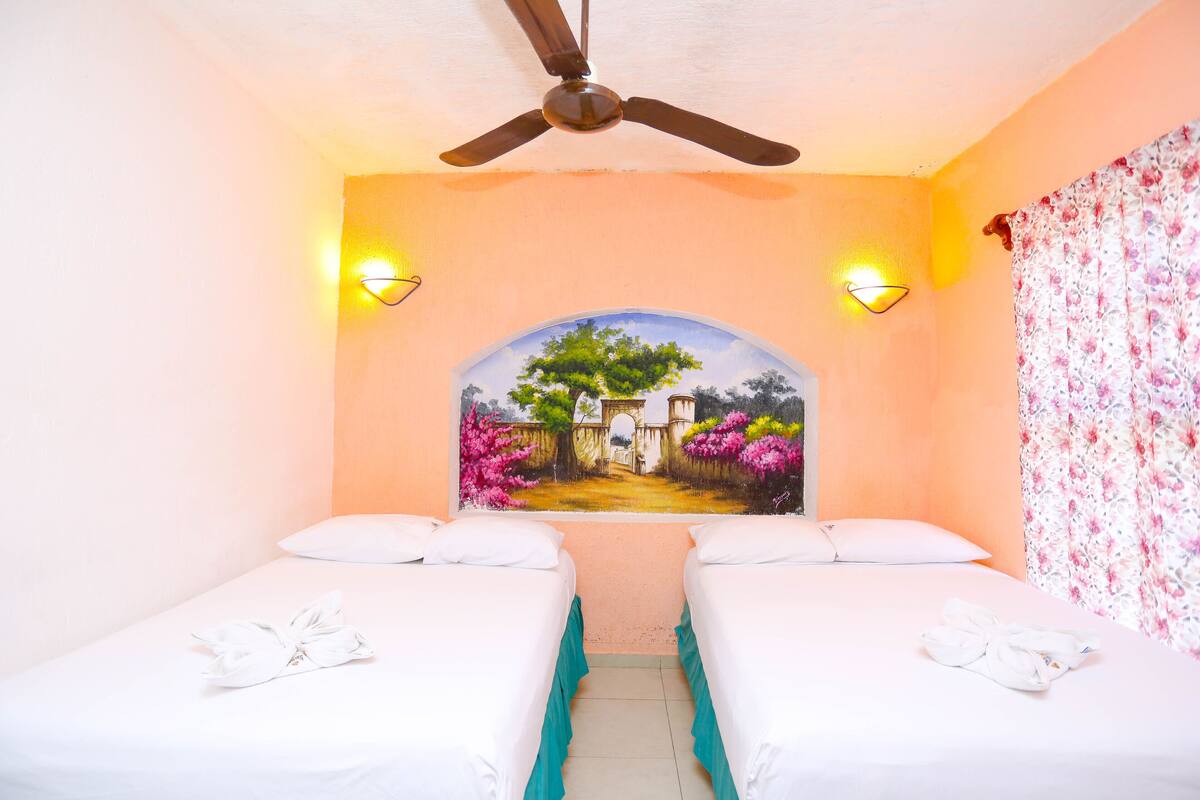 A hotel room with two beds, a ceiling fan, a painting of a garden, and a floral curtain.