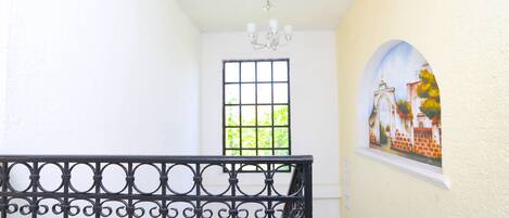 Un pasillo con barandilla de metal negro, un candelabro, una ventana con vistas a la vegetación y un cuadro en la pared.