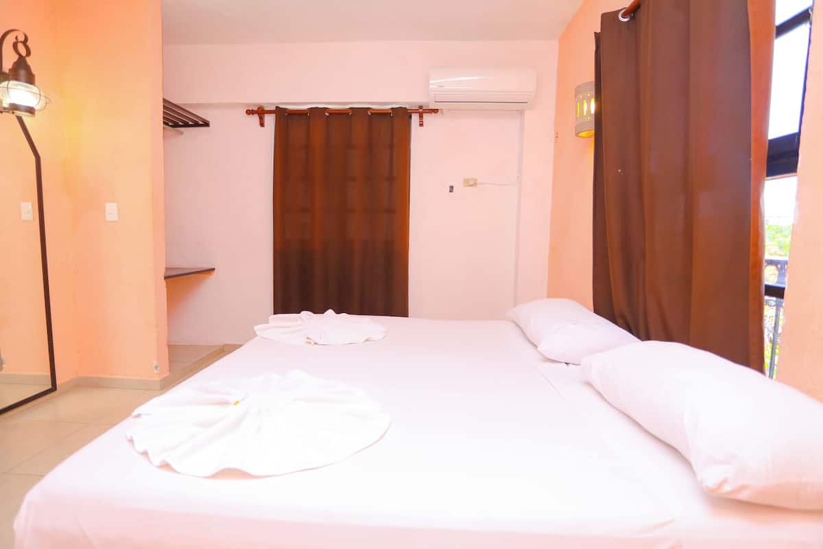A hotel room with a bed, a mirror, an air conditioning unit, and a window with curtains.
