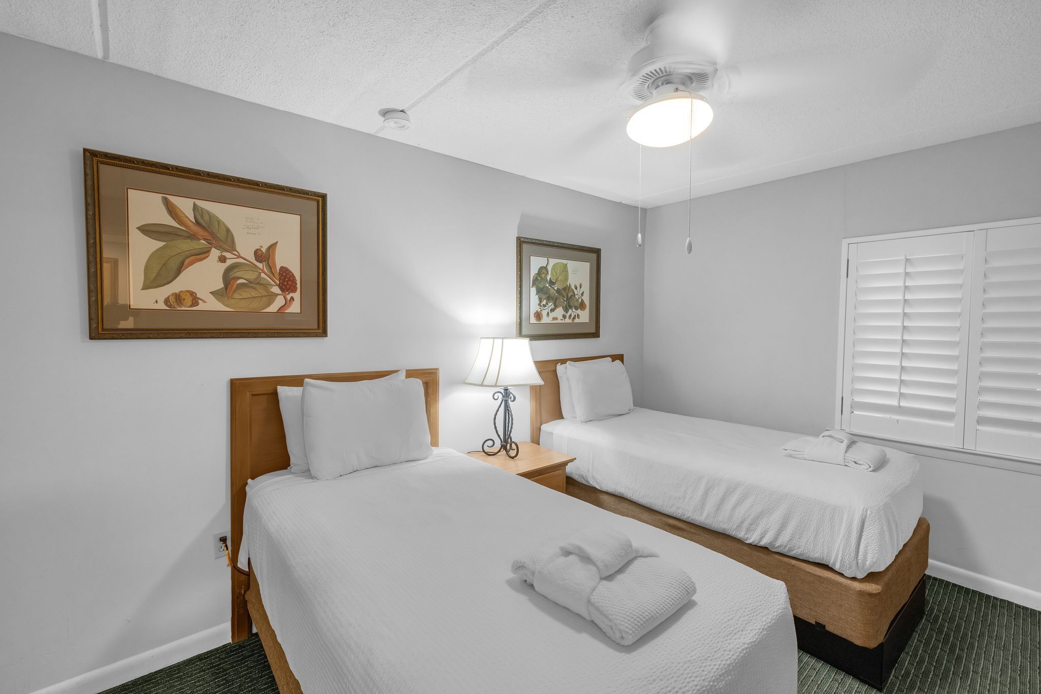 A hotel room with two single beds, a ceiling fan, a framed botanical print on the wall, and a window with blinds.