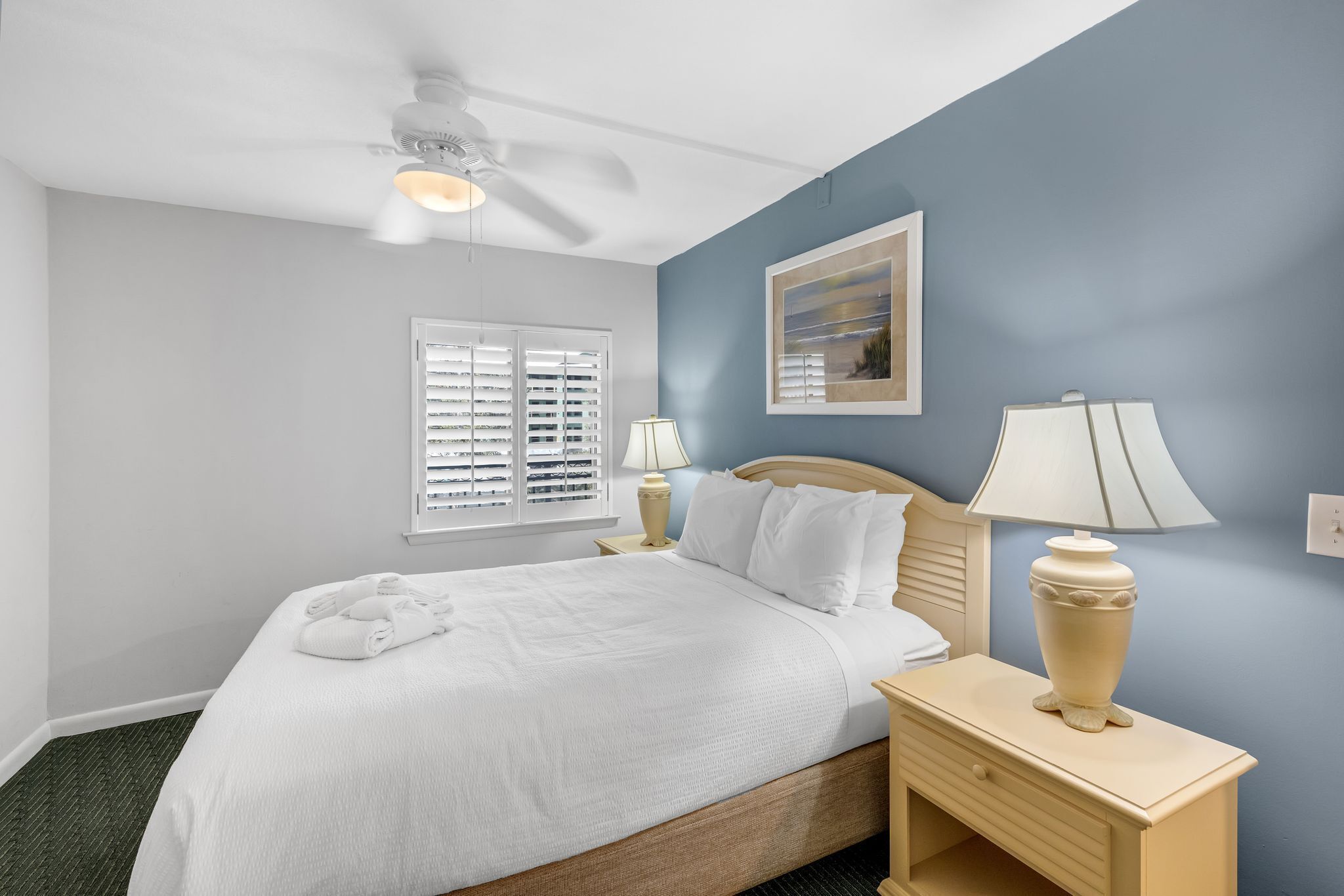 A neatly made bed with white linens, a bedside lamp, and a ceiling fan in a room with blue walls and a window with blinds.