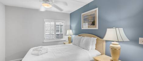 A neatly made bed with white linens, a bedside lamp, and a ceiling fan in a room with blue walls and a window with blinds.