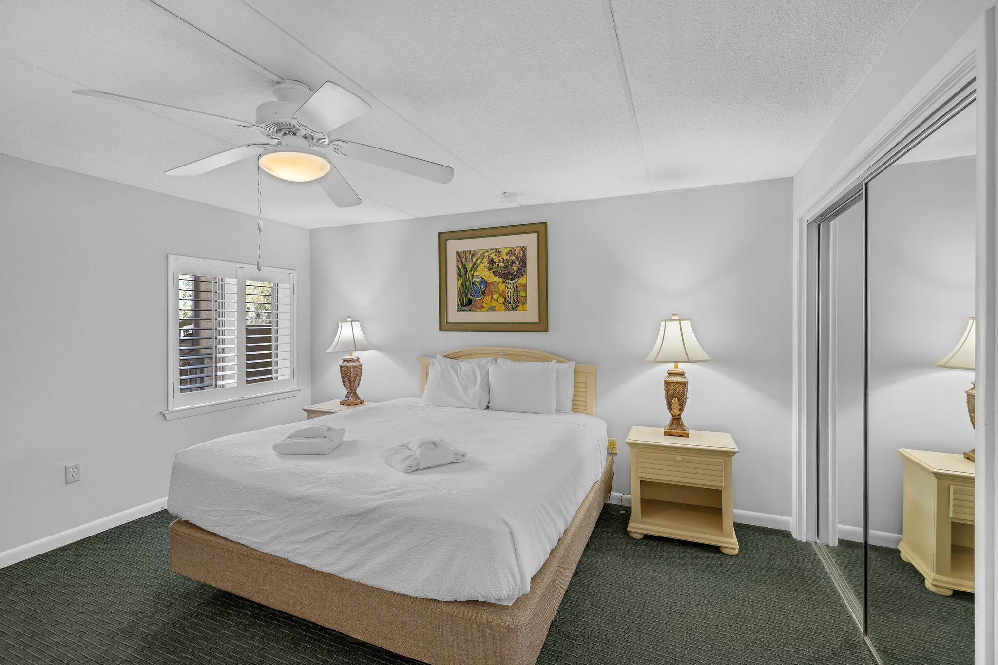 Una habitación de hotel con una cama grande, dos mesitas de noche con lámparas, un ventilador de techo, una ventana con persianas y un cuadro en la pared.