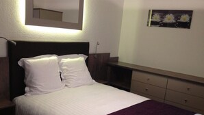 Una cama bien tendida con sábanas blancas y un detalle morado, una mesita de noche, un aparador con cajones y un espejo montado en la pared.