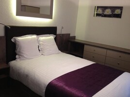 Una cama bien tendida con sábanas blancas y un detalle morado, una mesita de noche, un aparador con cajones y un espejo montado en la pared.