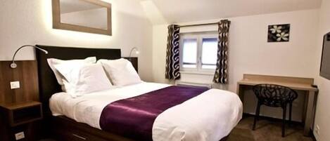 A hotel room with a bed, bedside table, desk, chair, and a window with curtains.