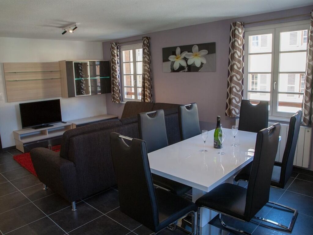 Een moderne woonkamer met een bank, eettafel en stoelen. Er staat een televisie op een dressoir en een kast met glazen deuren.