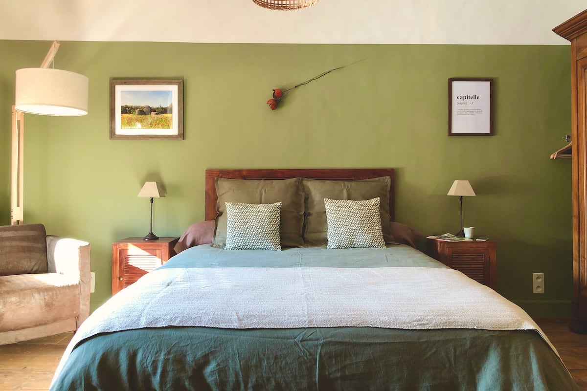 Un dormitorio con una cama, dos mesitas de noche, una lámpara, una silla y un cuadro en la pared.