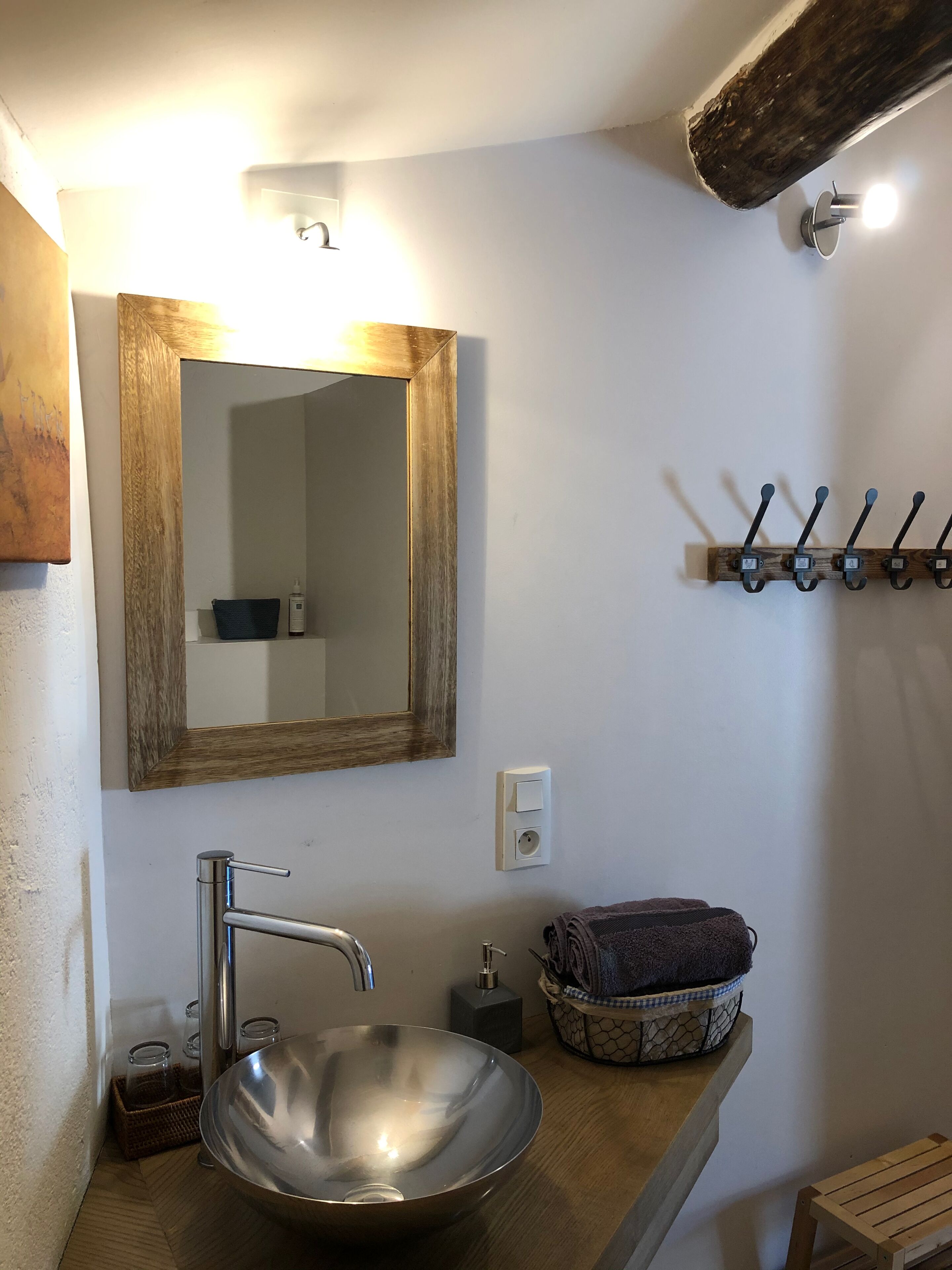 Casa de banho com um espelho grande, uma pia de aço inoxidável, uma prateleira de madeira com uma cesta e um porta-toalhas fixado na parede.