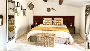 Een slaapkamer met een houten hoofdbord, een groot bed met gekleurde kussens, een nachtkastje met een lamp, en een geweven bankje.