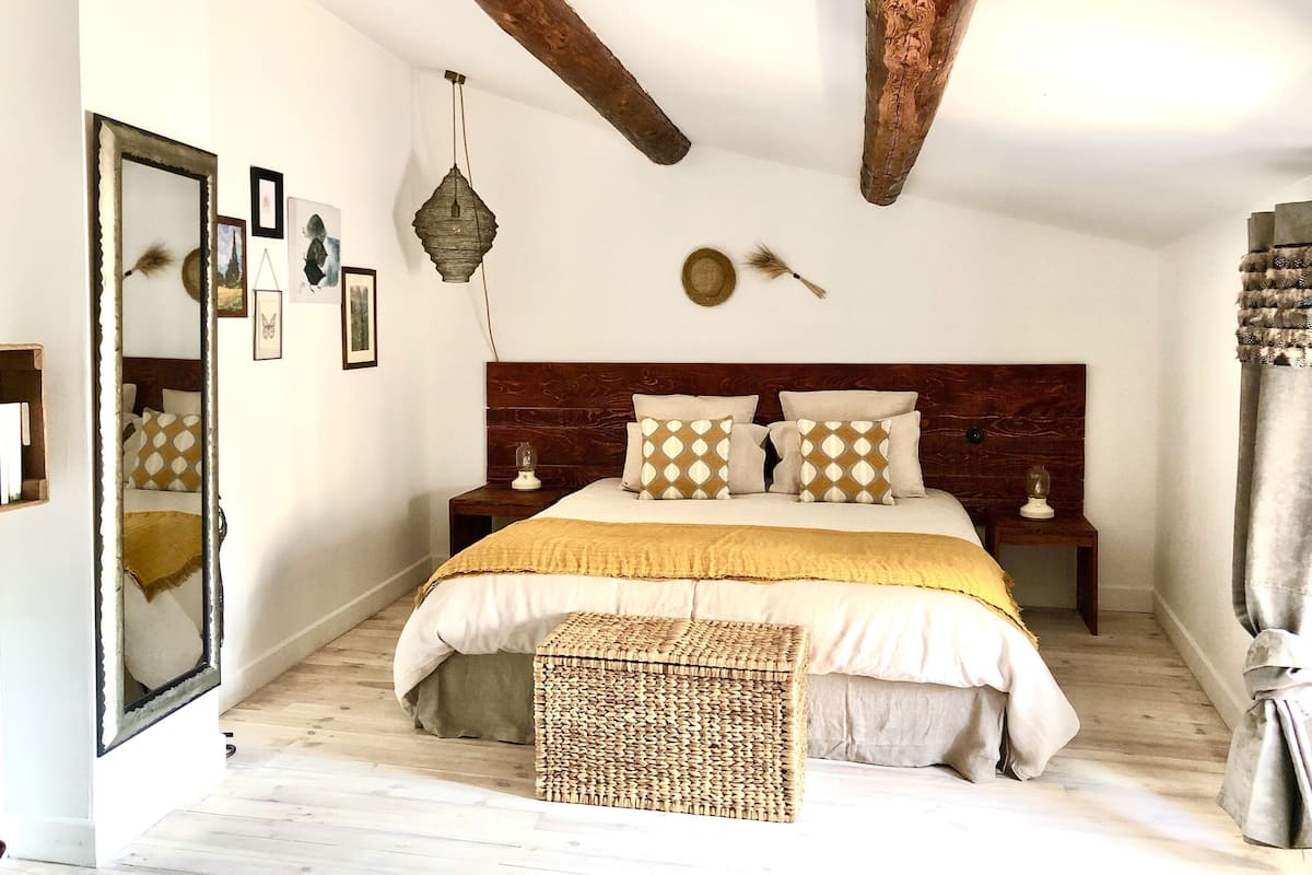 Un dormitorio con cabecera de madera, una cama grande con almohadas estampadas, una mesita de noche con lámpara y un banco tejido.