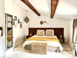 Un dormitorio con cabecera de madera, una cama grande con almohadas estampadas, una mesita de noche con lámpara y un banco tejido.