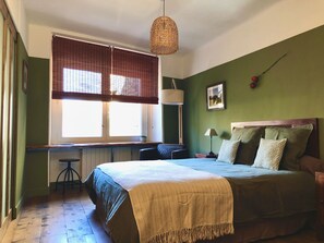 Een slaapkamer met een bed, een bureau, een stoel, een lamp en een raam met jaloezieën.