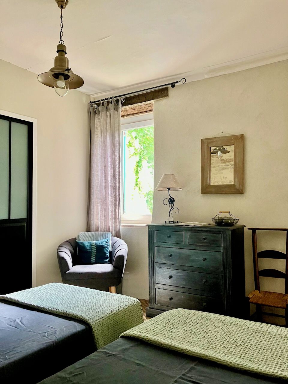 A room with two beds, a chair, a dresser, a lamp, and a window with curtains.