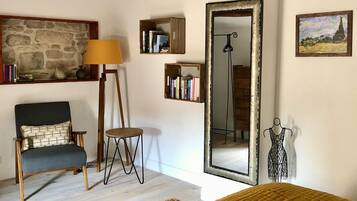 A room with exposed wooden beams, a stone wall, a mirror, a mannequin, and a bed with a mustard-colored blanket.
