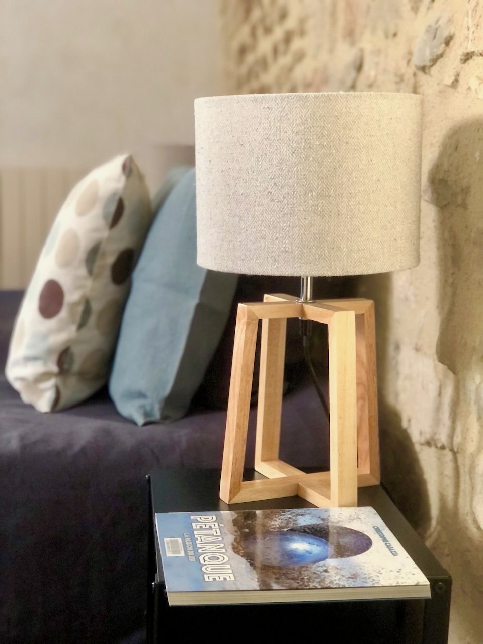 A wooden floor lamp with a fabric shade on a black table, a book titled 'PETANCHE' beside it, and a patterned pillow on a couch in the background.