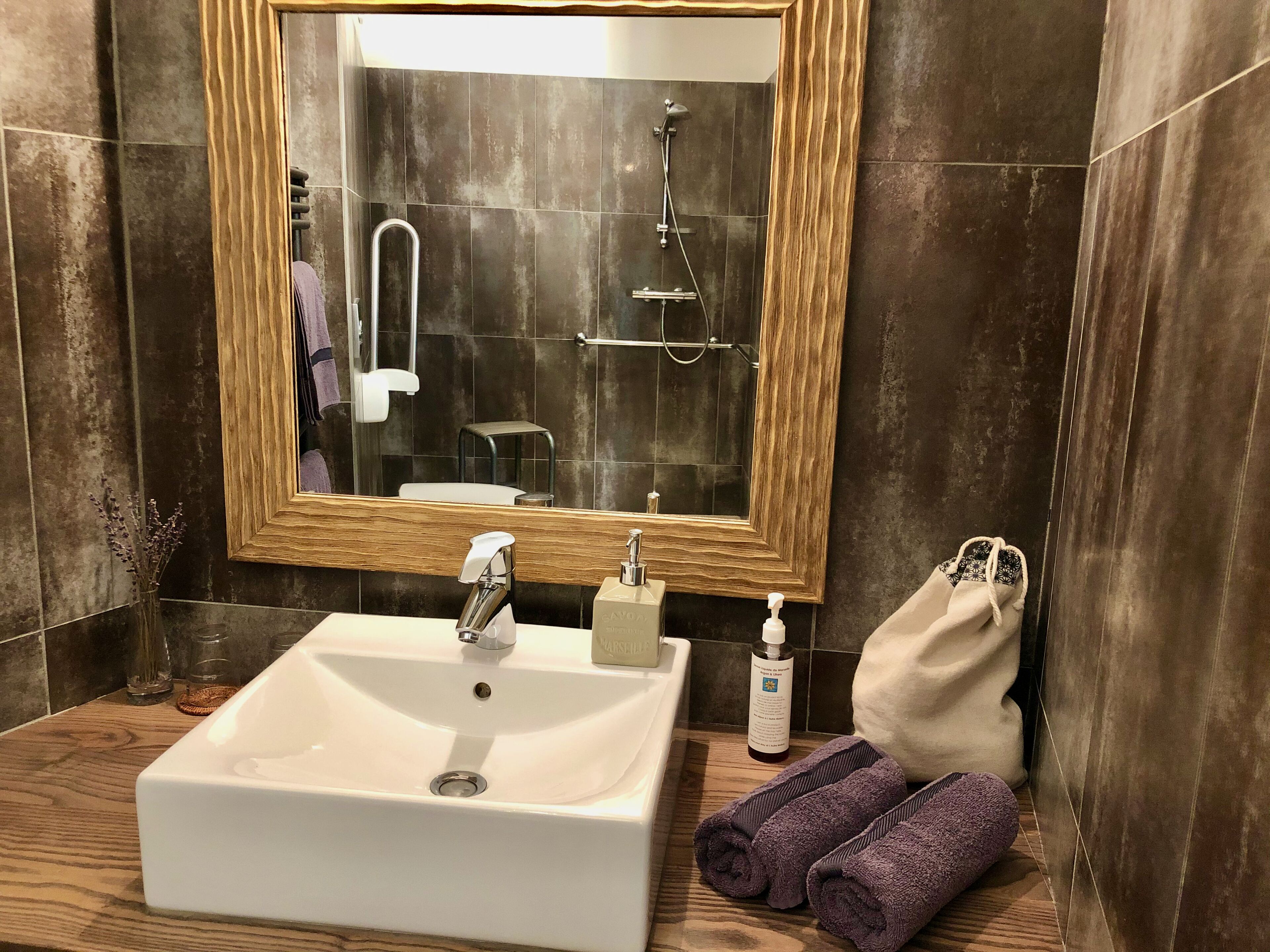 Un baño con un espejo de marco de madera, un lavabo blanco, un cabezal de ducha de mano, una toalla y una botella de jabón.