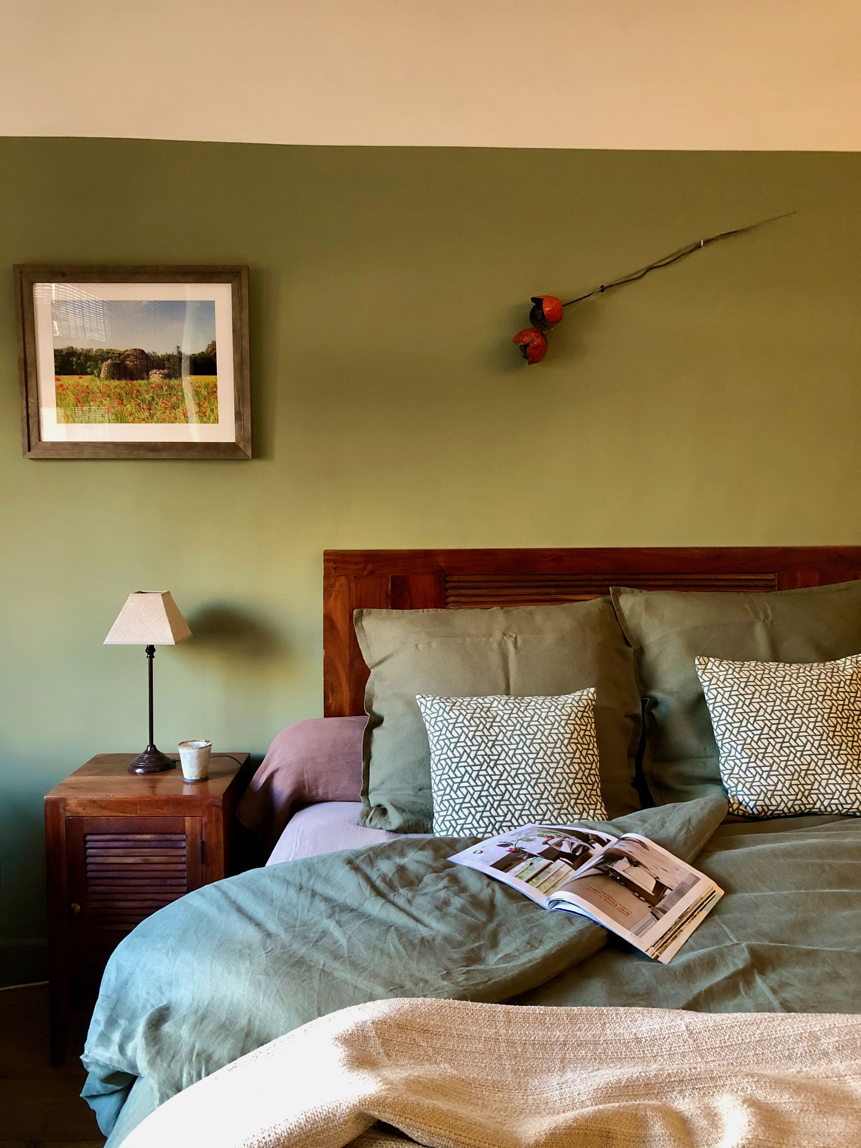 A neatly made bed with a magazine, a lamp, and a framed picture on a nightstand.