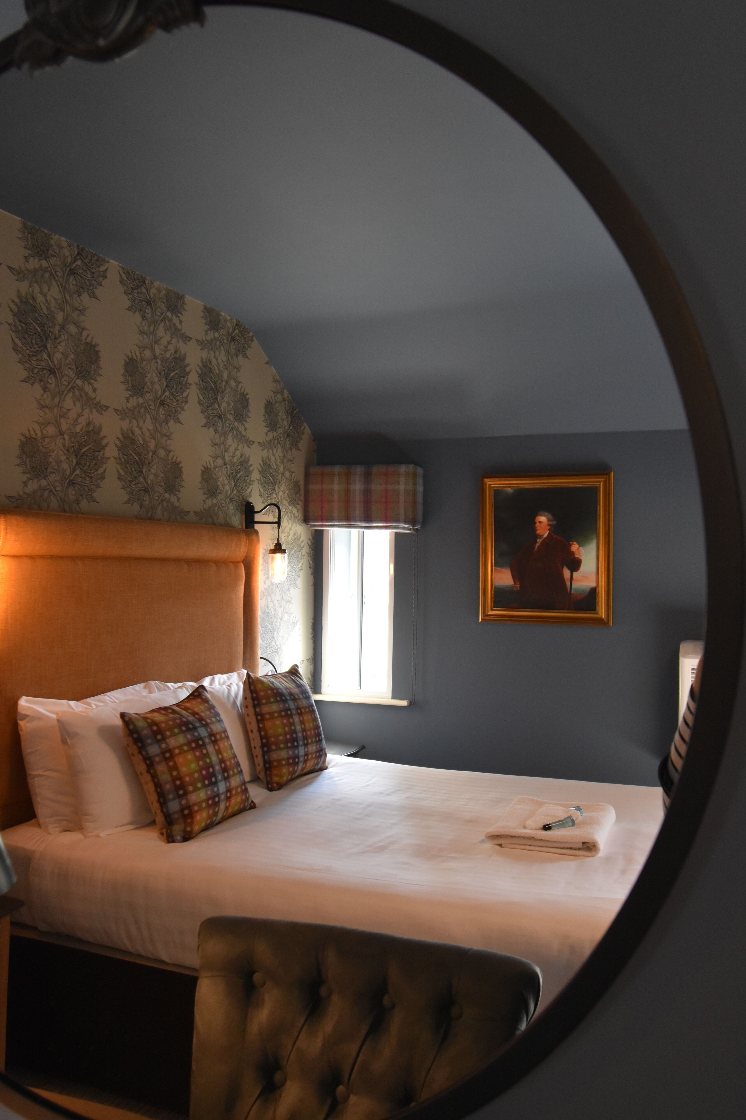 Ein Schlafzimmer mit einem großen Bett, einem Kopfteil, einem Fenster mit einem Karomuster-Vorhang, einem gerahmten Bild an der Wand und einem runden Spiegel.