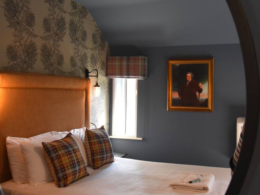 A bedroom with a large bed, a headboard, a window with a plaid curtain, a framed picture on the wall, and a round mirror on the wall.