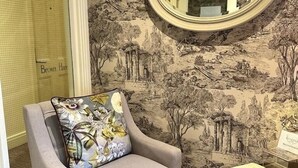 Ein Zimmer mit einem grauen Sessel, einem blumigen Kissen, einem Spiegel und einer Wand mit naturbezogenen Tapeten.