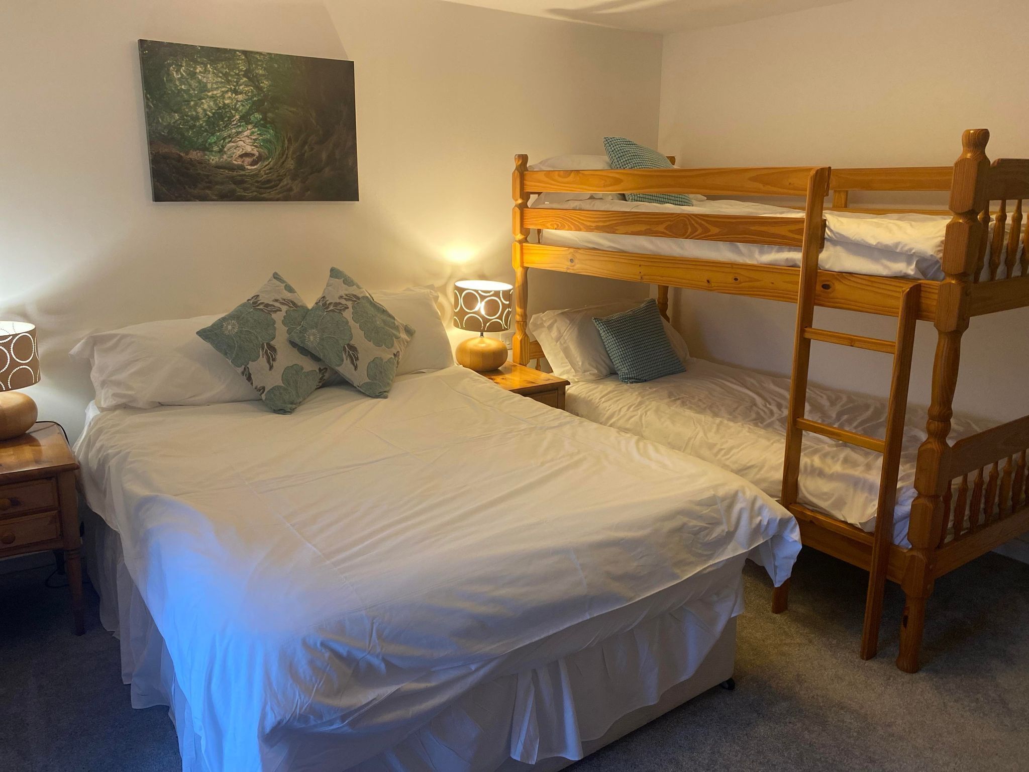 Quarto com duas camas, uma com estrutura de madeira e outra com estrutura de metal. Há uma pintura na parede, um abajur na mesa de cabeceira e uma janela com cortinas.