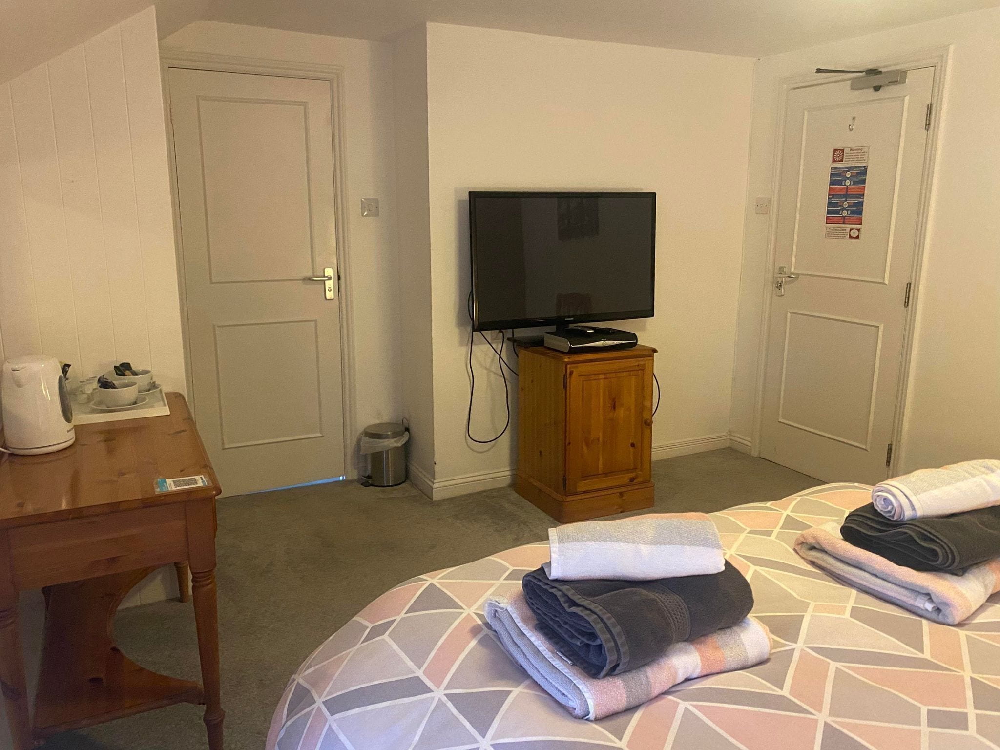 Habitación de hotel con una cama, un televisor sobre un mueble de madera, una mesita con una tetera y un cesto de basura.