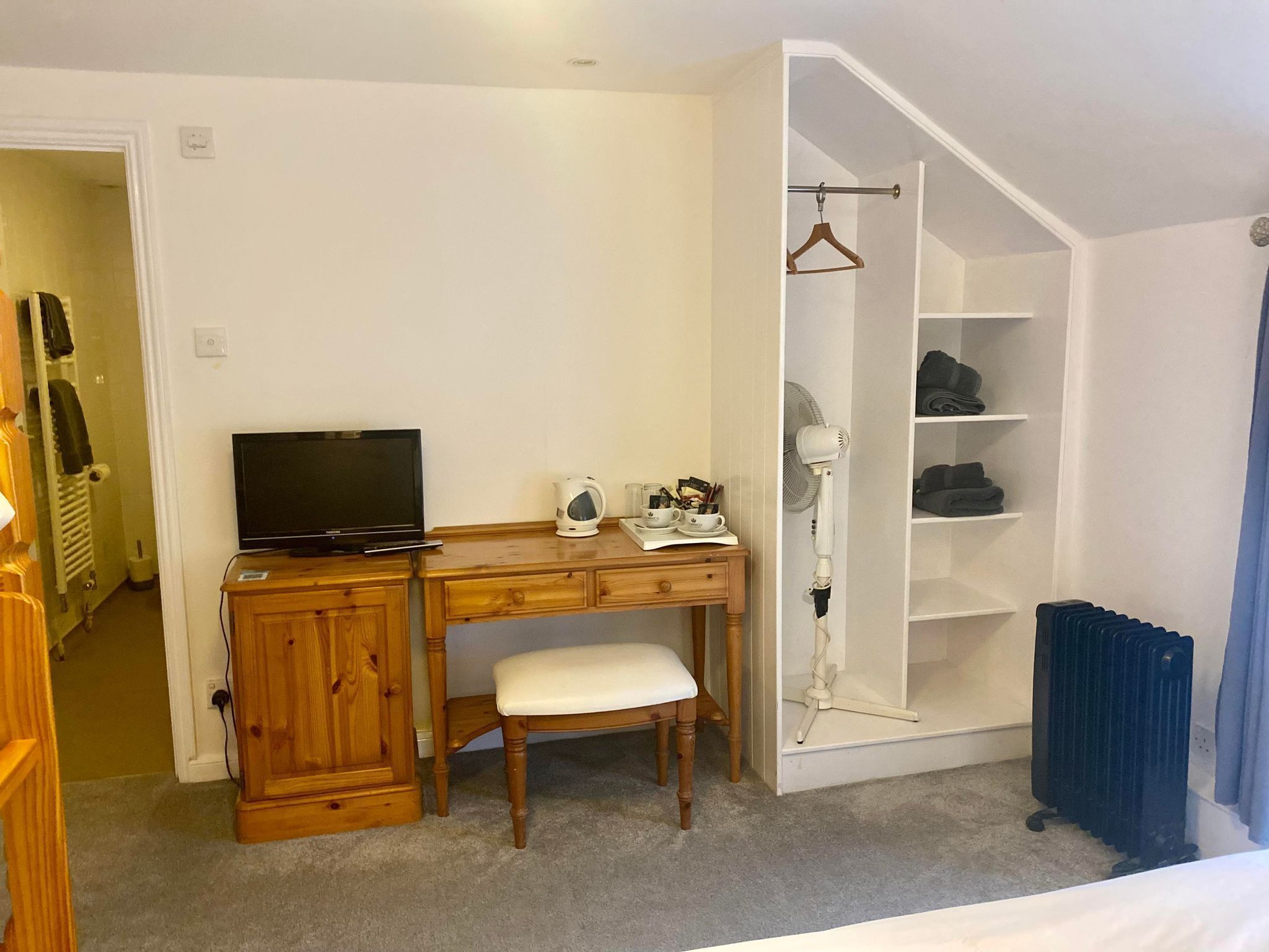 Une chambre avec un bureau en bois, une télévision, une table de chevet avec une lampe, une chaise et un dressing avec des étagères.