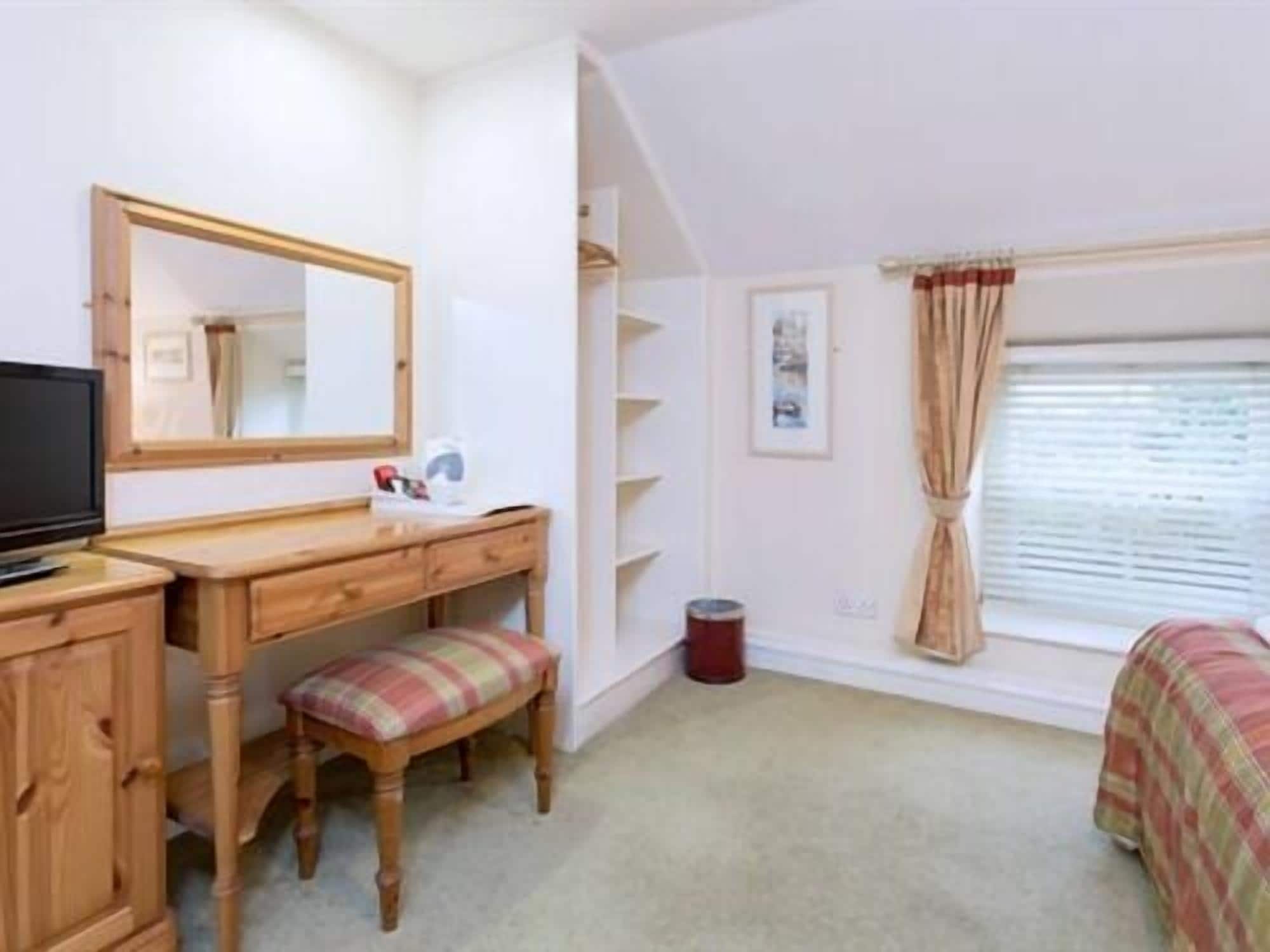 A bedroom with a wooden desk, a chair, a television, a window with curtains, and a bed with a plaid bedspread.