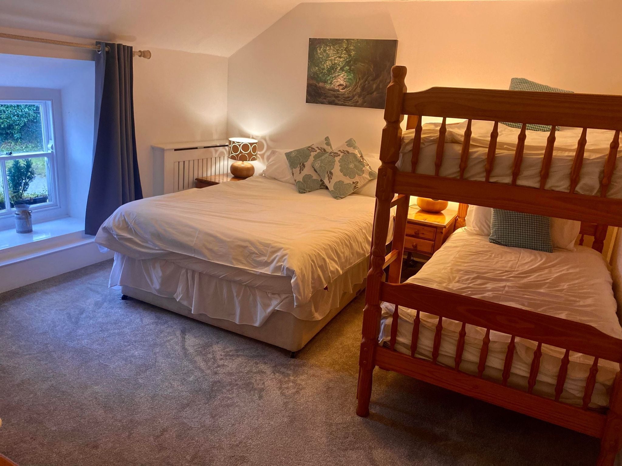 A bedroom with a large bed, a smaller bed, a nightstand with a lamp, a window with curtains, and a painting on the wall.