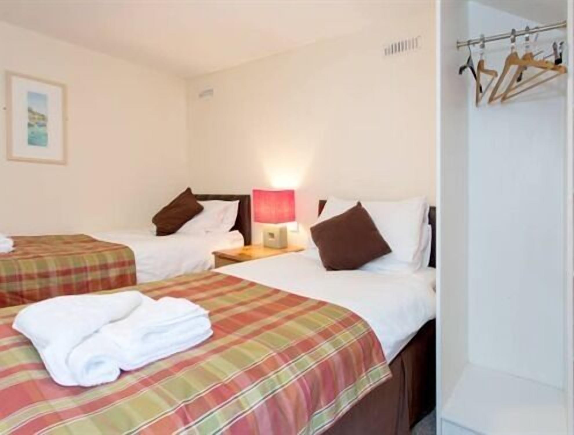A hotel room with two beds, each with a plaid bedspread, a white towel on the first bed, and a nightstand with a lamp.