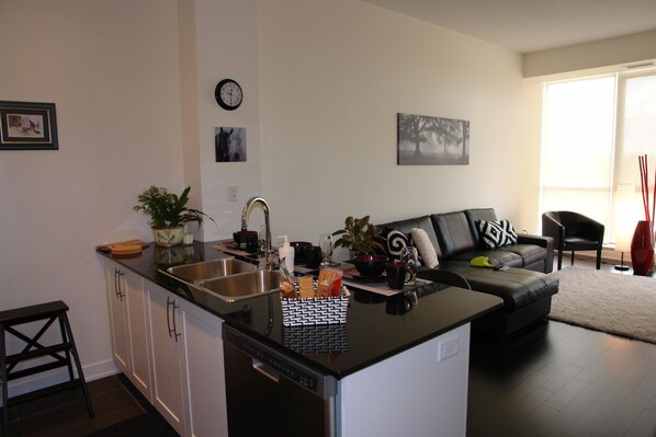 A modern kitchen with a black countertop, white cabinets, and a built-in dishwasher. There is a dining area with a black sofa and a coffee table. - Goodwood Suites (Mississauga)