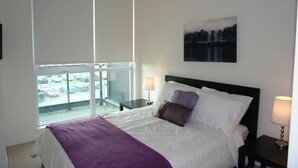 Una habitación de hotel con una cama, una ventana que da a coches aparcados, un cuadro en la pared y una mesita de noche con una lámpara.
