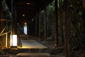 A covered wooden walkway with stone steps leading to a door, surrounded by bamboo and foliage. - Hana no kumo (Ito)