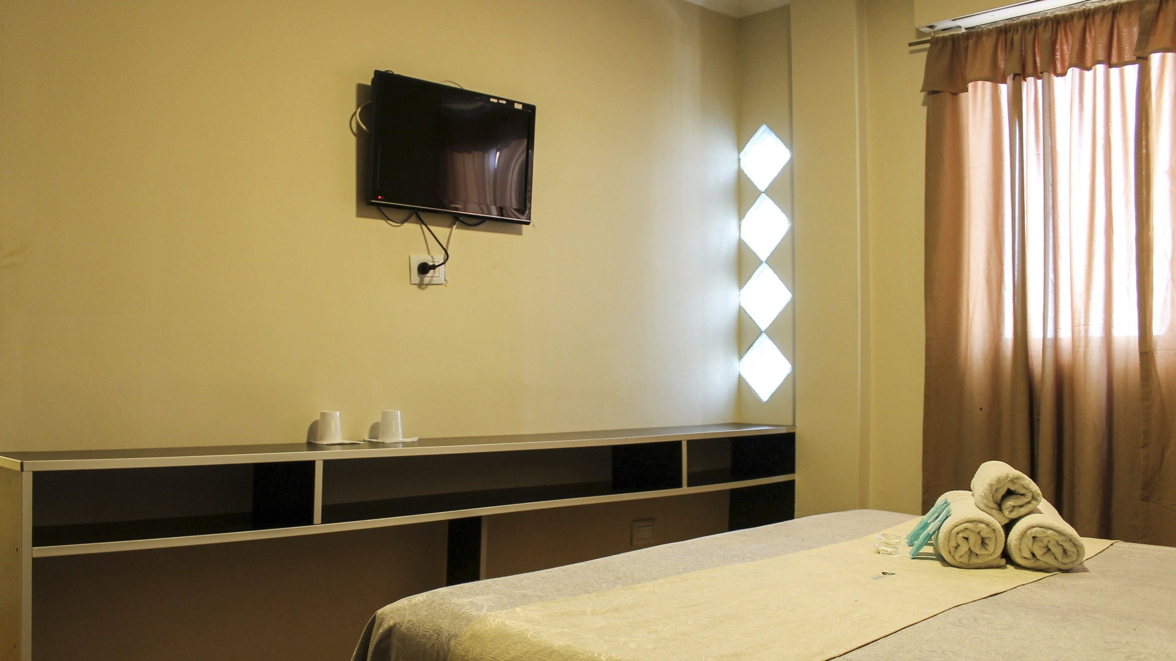 A hotel room with a bed, a flat-screen TV mounted on the wall, a window with curtains, and a small table with cups and towels.