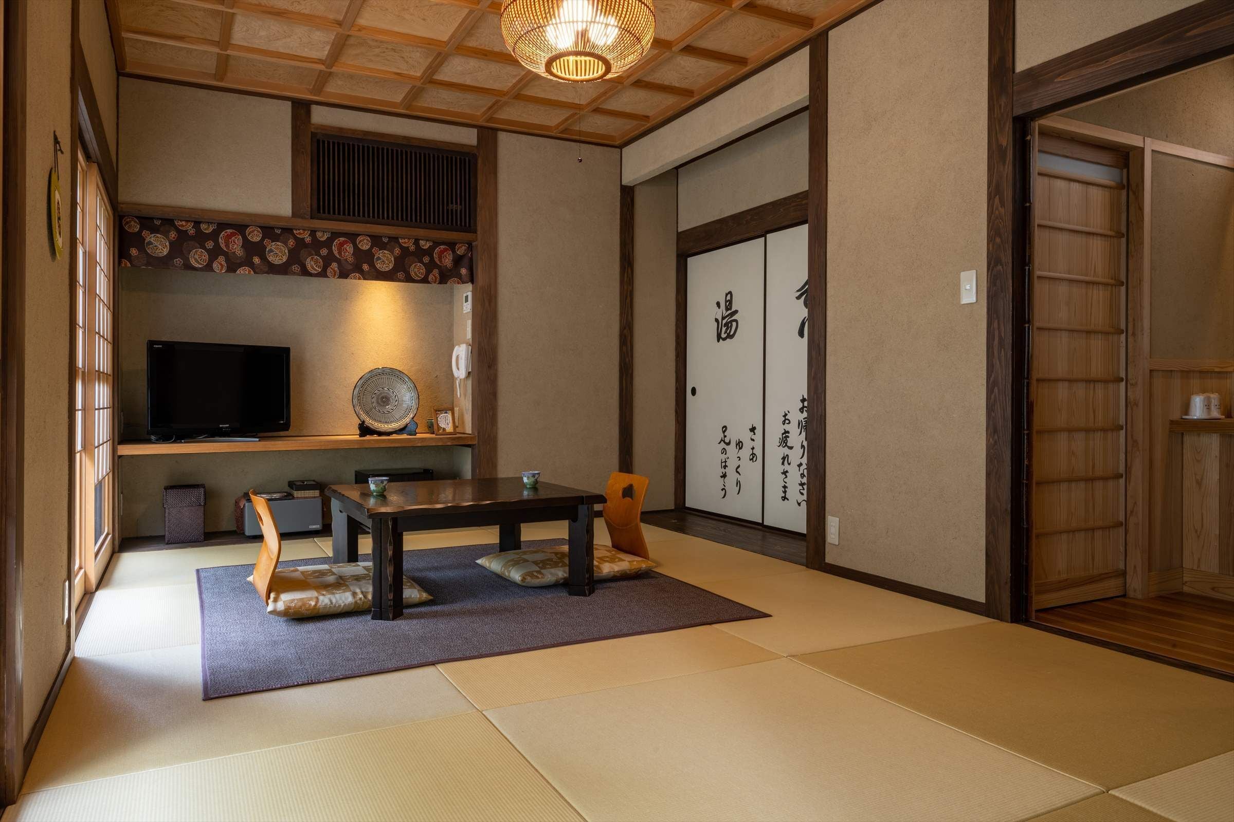 A traditional Japanese room with tatami flooring, a low table with cushions, a television, and sliding doors with calligraphy.