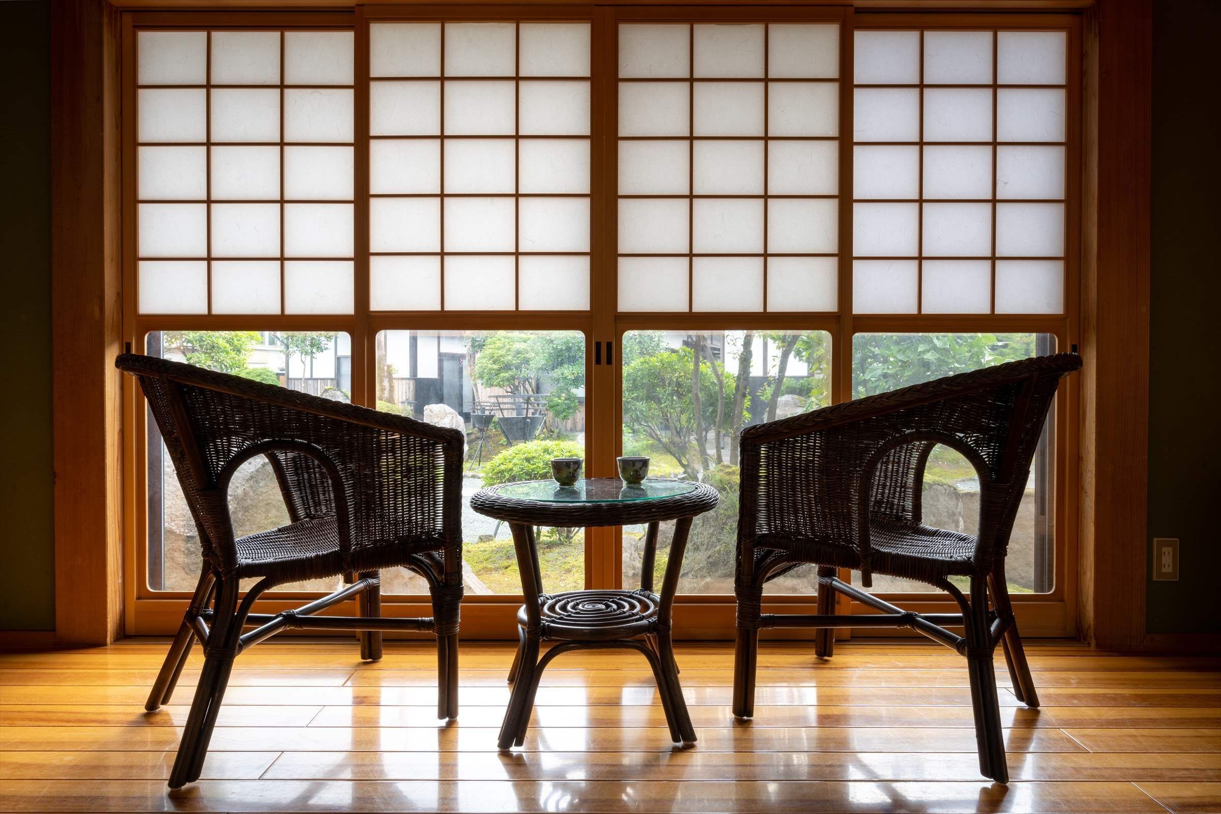 Quarto com duas cadeiras de vime, uma pequena mesa redonda, uma grande janela com tela shoji e piso de madeira.