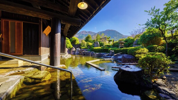 Un onsen japonés tradicional con estructura de madera, un sendero de piedra y un pequeño estanque con rocas y plantas.