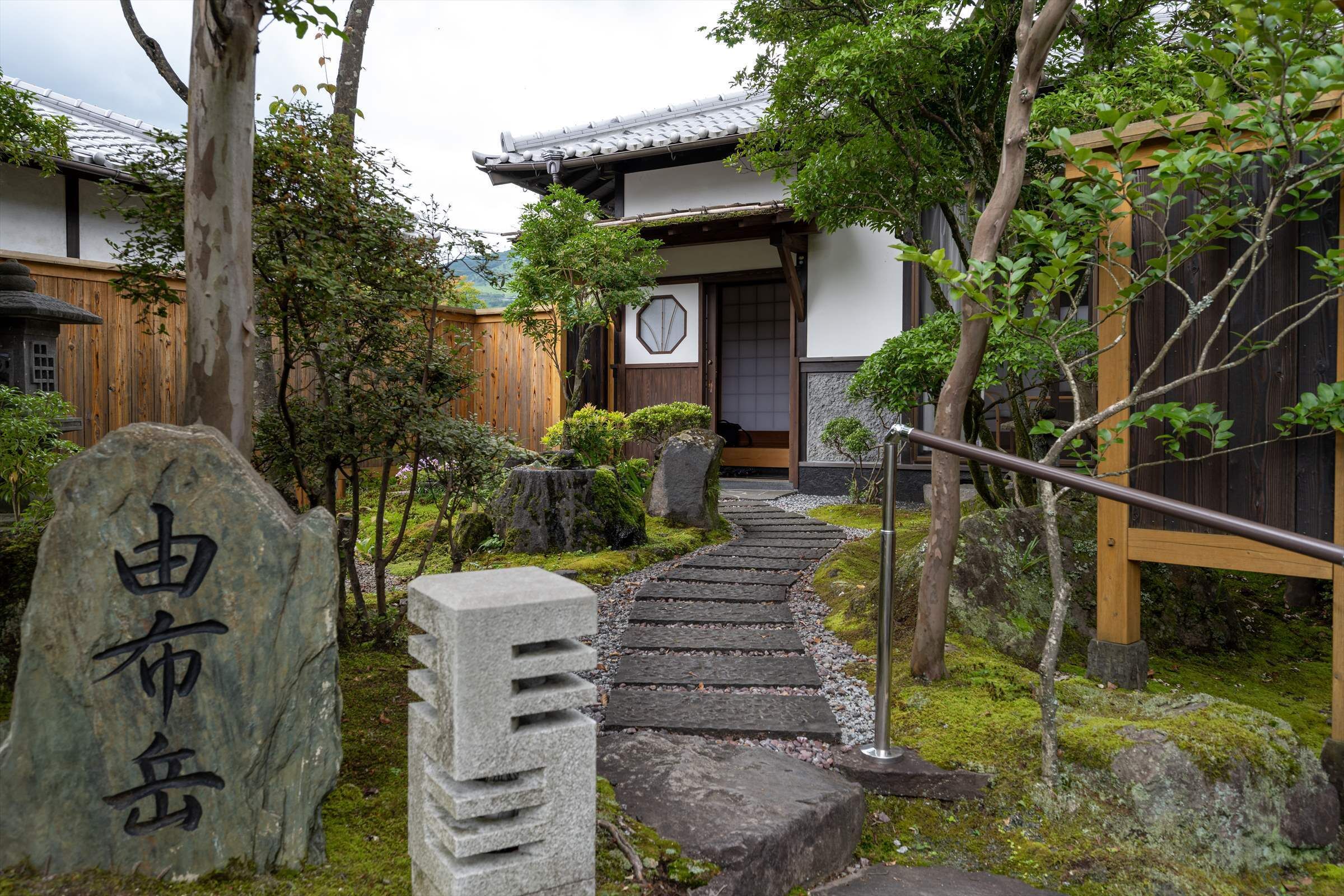 A traditional Japanese building with a wooden exterior and a tiled roof. A stone pathway leads to the entrance, surrounded by greenery and rocks.