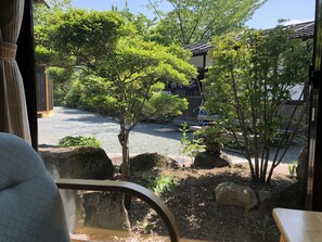 A view from a window with a view of a gravel path, trees, and a building with a traditional roof. - Makibanoie (Yufu)