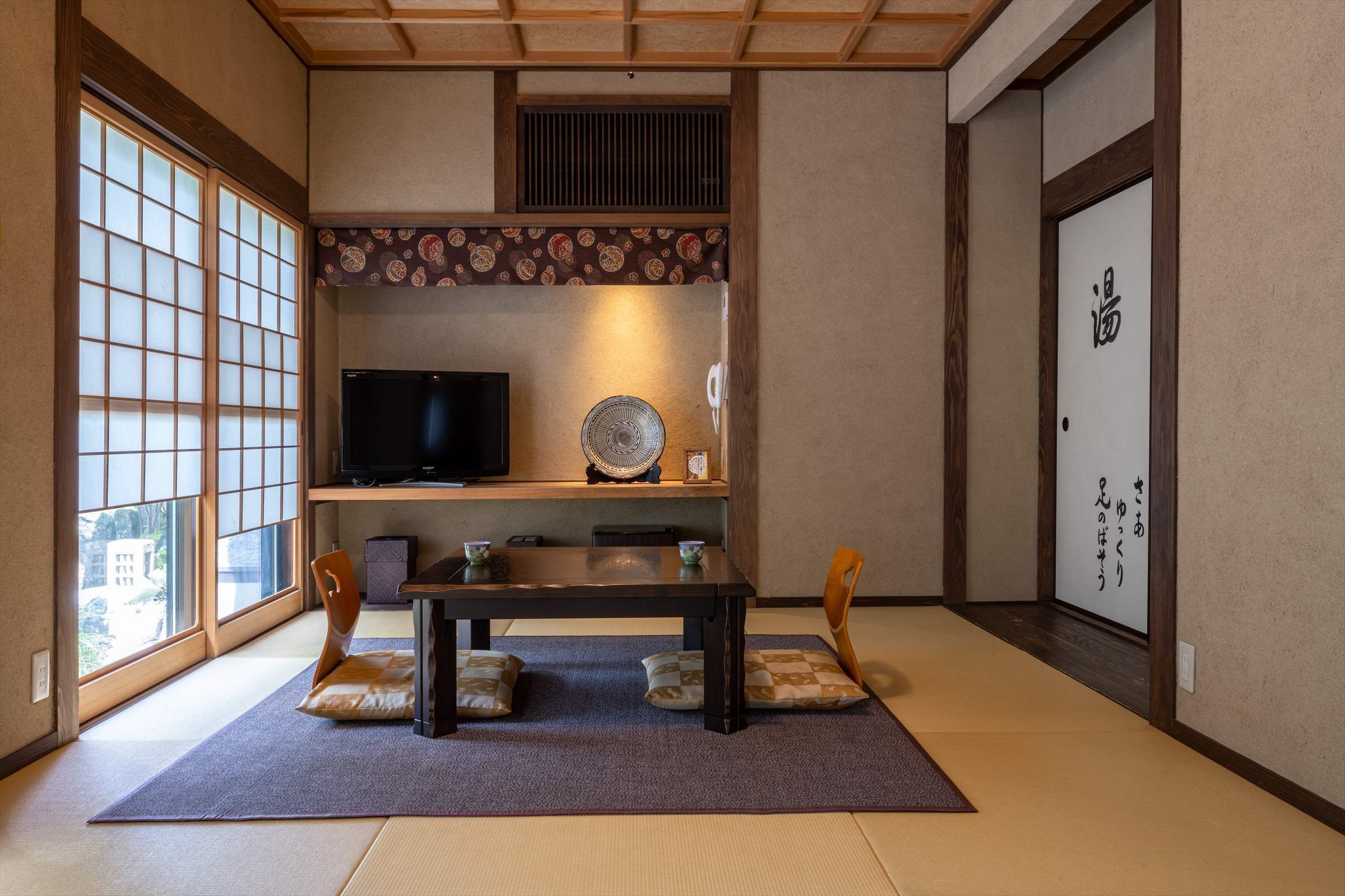 A traditional Japanese room with tatami flooring, a low table with cushions, a television, and a fan.