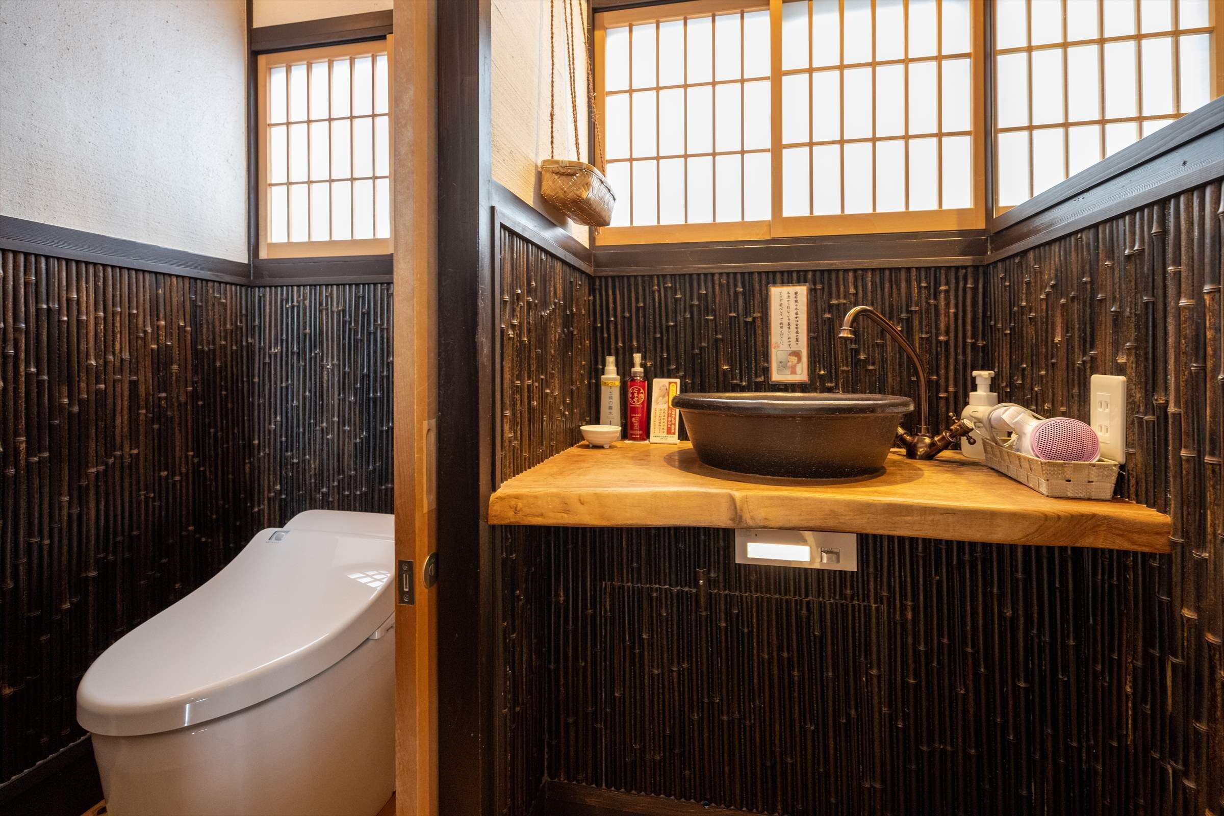 Ein traditionelles japanisches Badezimmer mit Bambuswand, Toilette, Waschbecken mit großer Schale und Fenster mit Gittermuster.