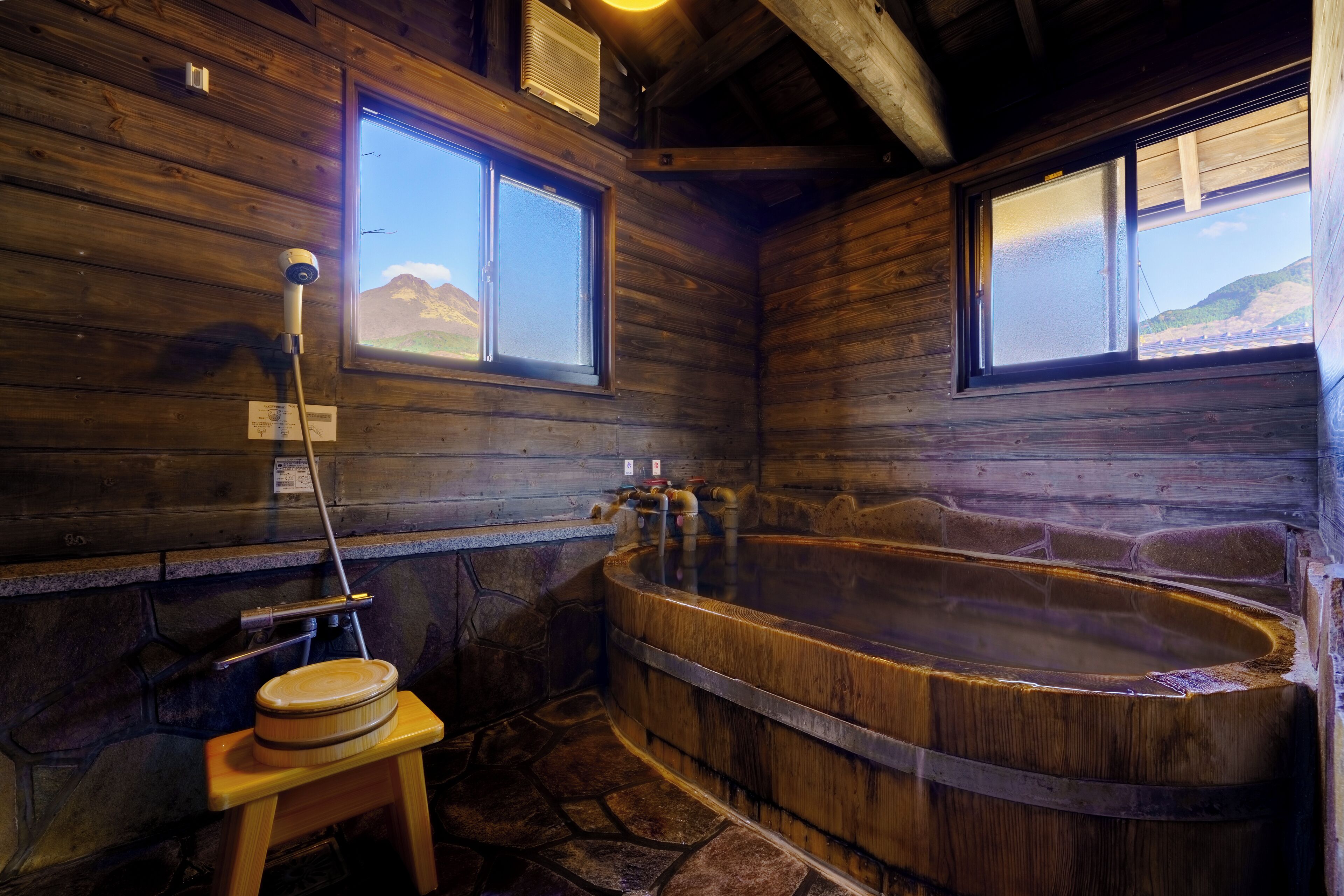 A wooden cabin with a large wooden bathtub, a small wooden stool, and a window with a view of mountains.