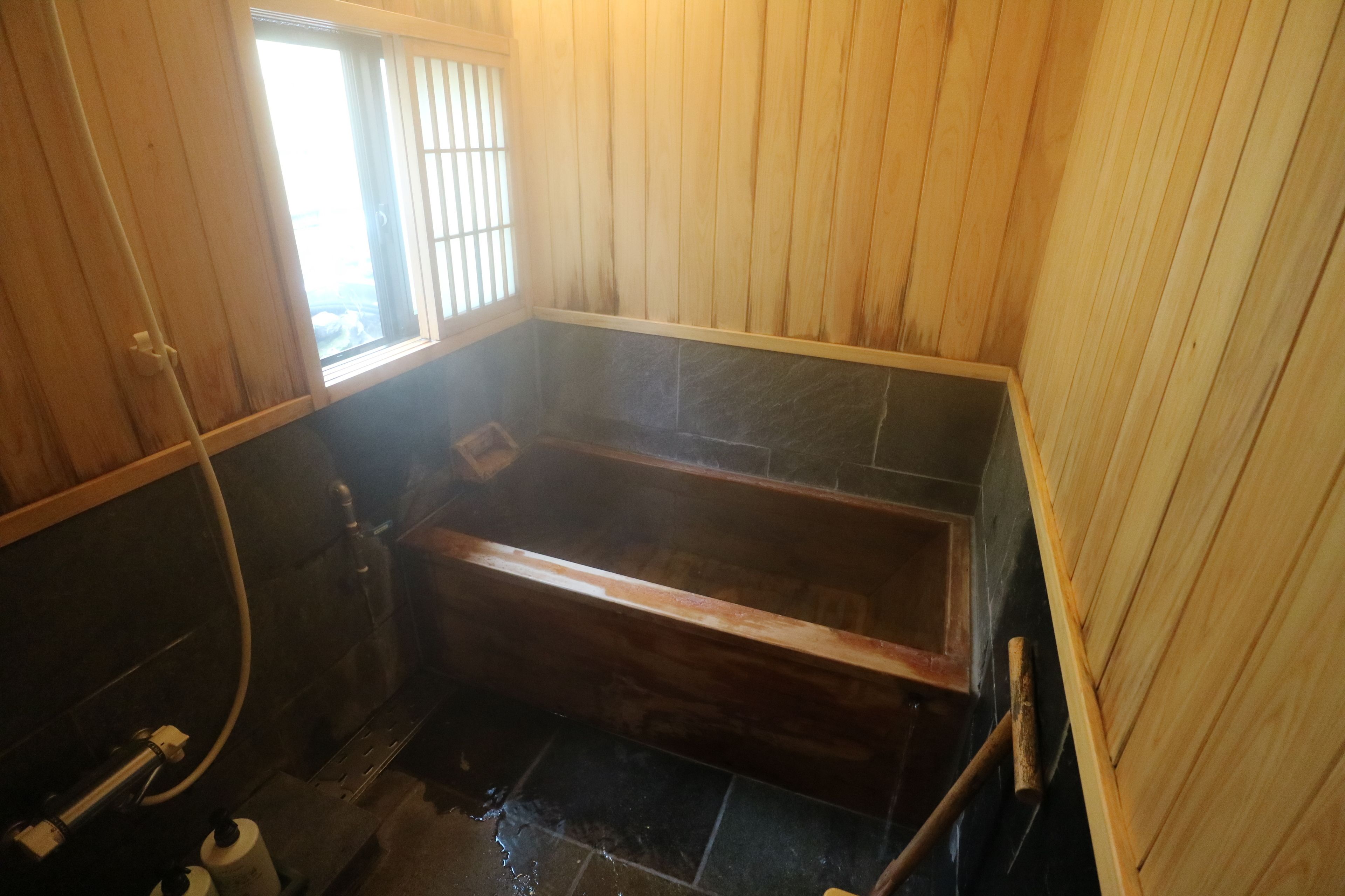 A wooden-paneled bathroom with a wooden bathtub, a window, and a shower area.