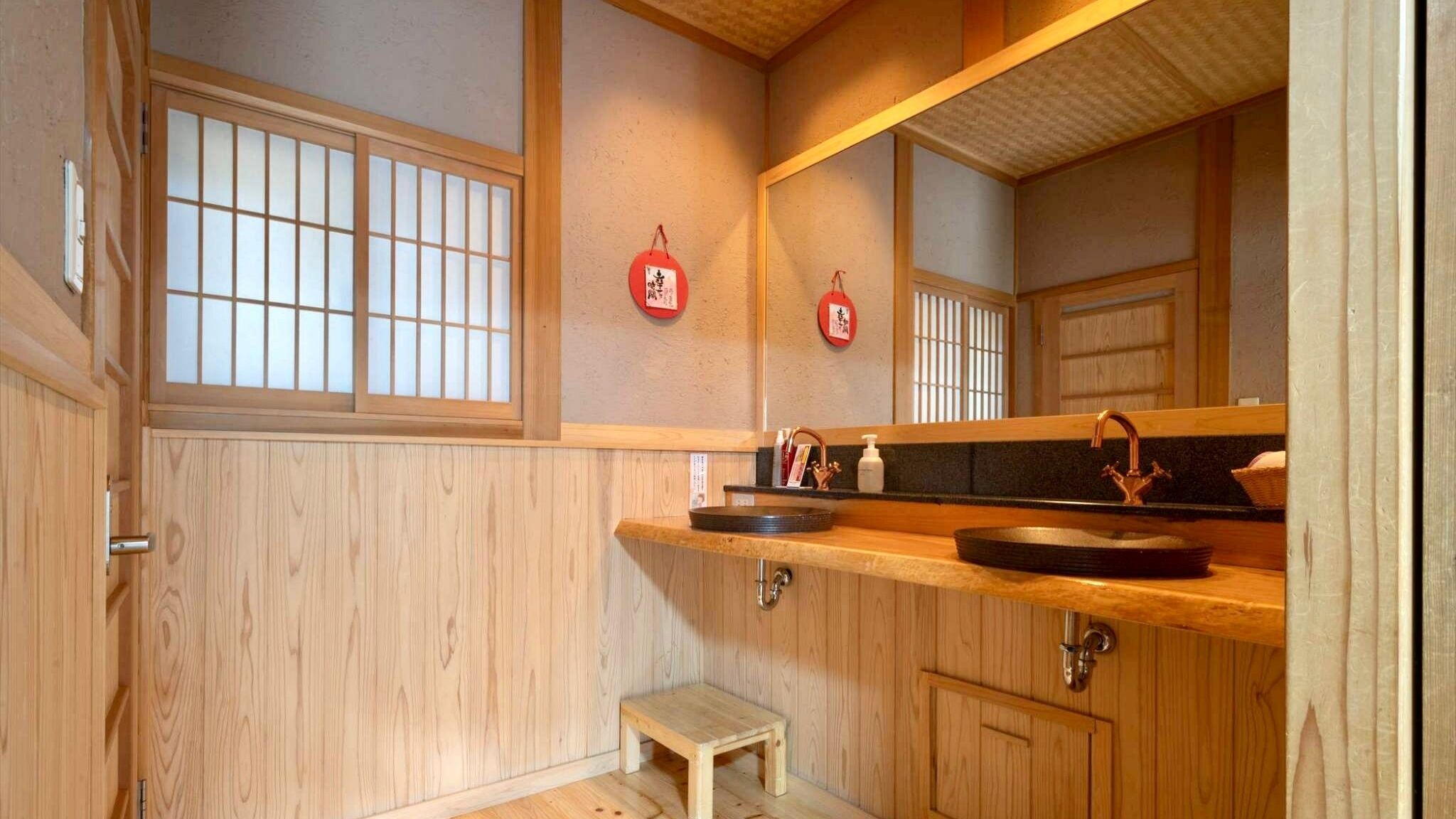A traditional Japanese-style bathroom with a wooden floor, a small wooden stool, a sink with a copper faucet, and a mirror above the sink.