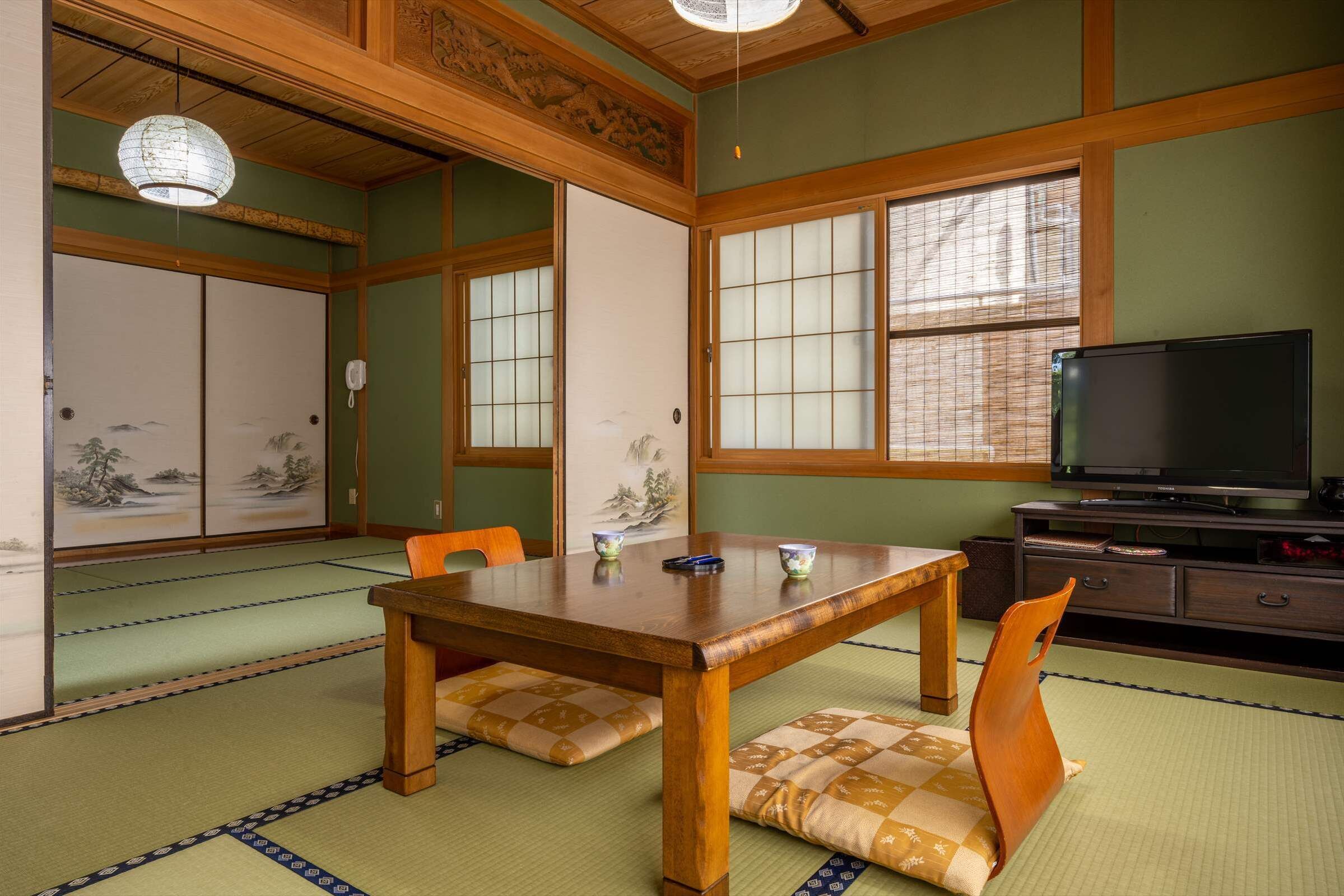 A traditional Japanese room with tatami flooring, a wooden table, chairs, a television, and sliding doors with artwork.